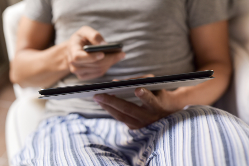 closeup of a young caucasian man in pajamas sitting in a comfortable armchair working from home using a smartphone and a digital tablet