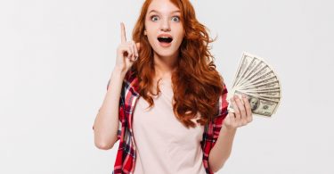 Picture of excited pretty young redhead lady standing isolated over white wall background looking camera while holding money have an idea.